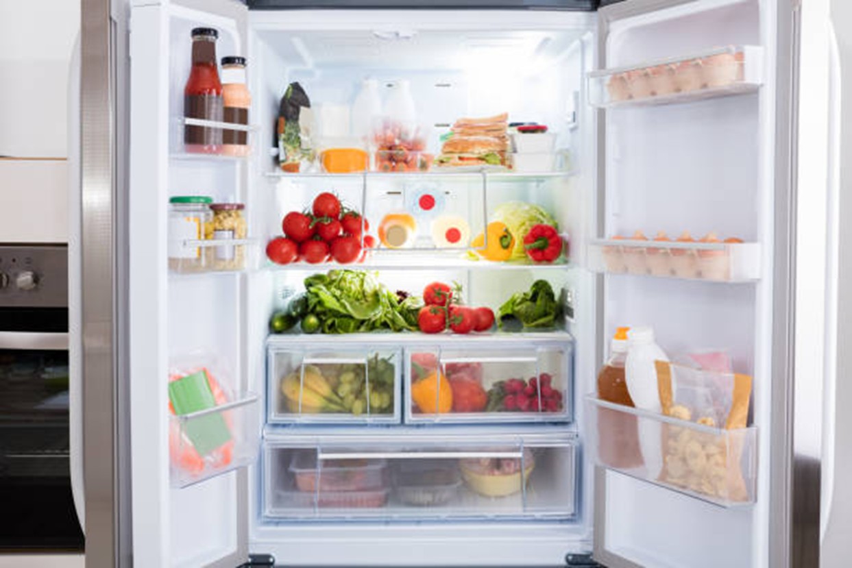An open, well-organized double-door refrigerator filled with fresh groceries. The shelves contain various vegetables like tomatoes, peppers, and lettuce, alongside eggs, milk, yogurt, and prepared containers. The lighting is bright, highlighting a clean and stocked kitchen interior.