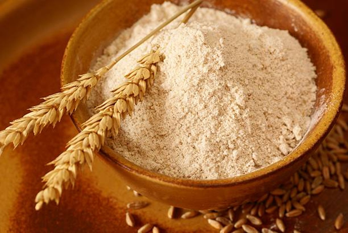 A rustic ceramic bowl filled with fine, tan-colored whole grain flour. Two stalks of dried wheat lie across the top of the flour. The bowl sits on a warm-toned surface scattered with loose wheat kernels